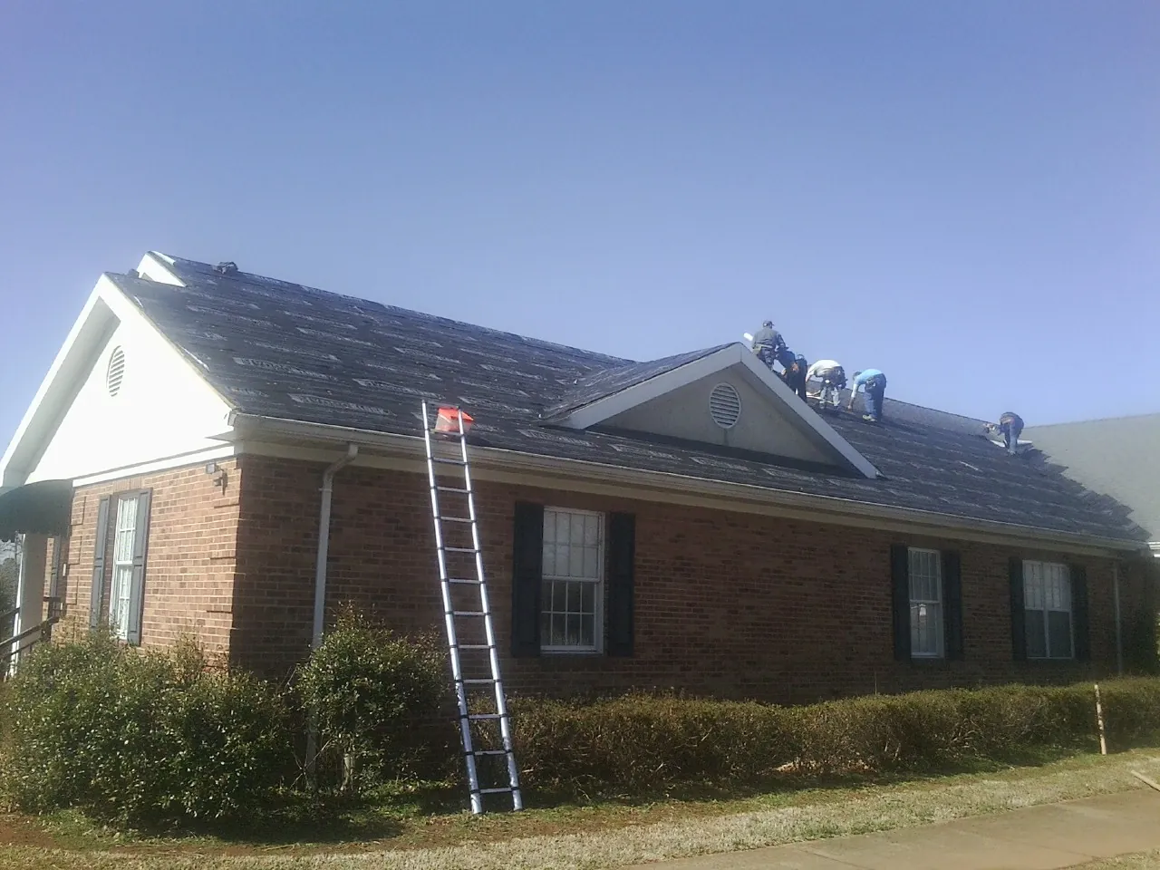 Crew tearing shingles off a brick ranch roof with a ladder set — Emergency Roof Repair work by our crew in West Granby