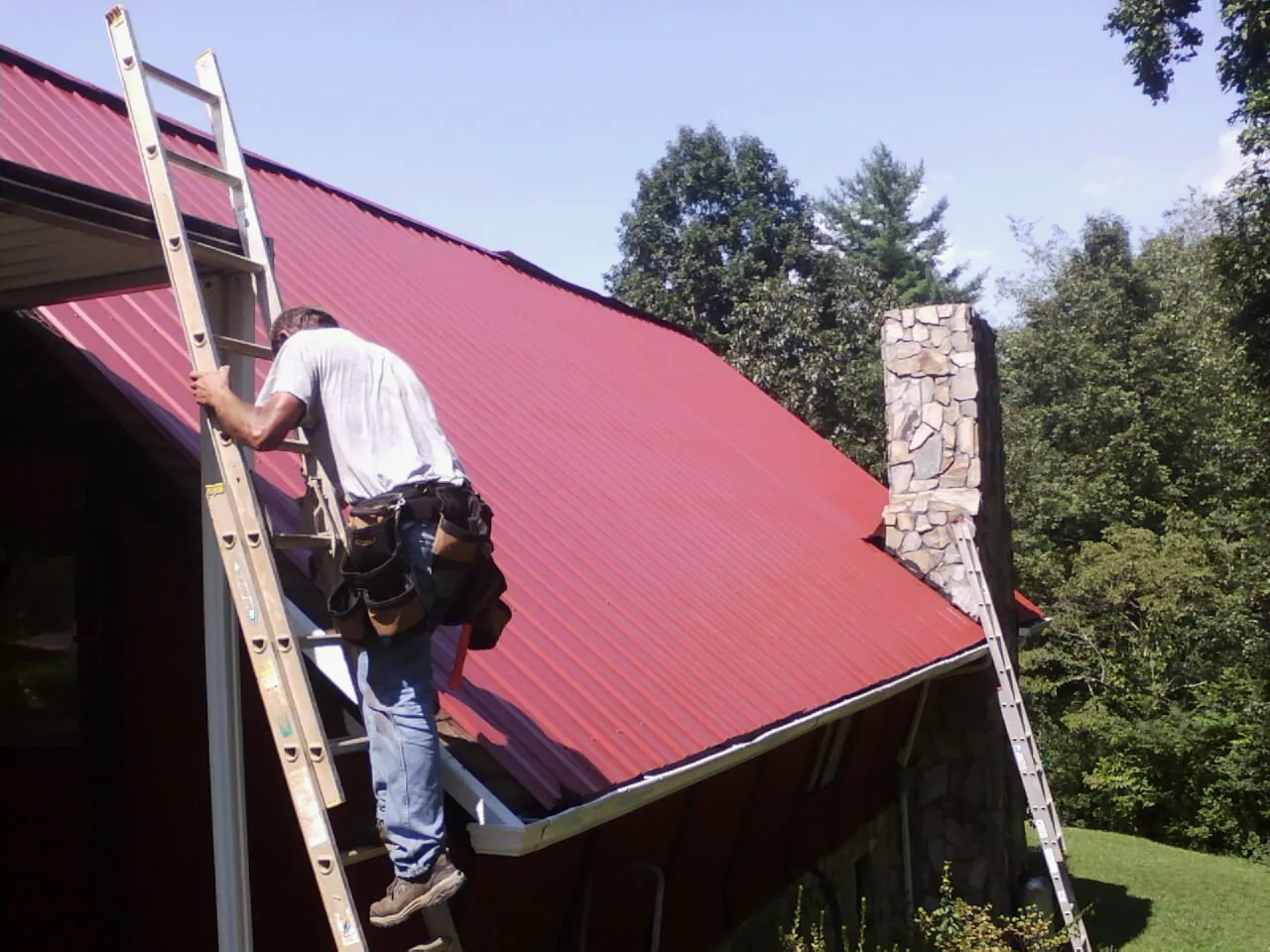 Roofer on a ladder beside a red metal roof and stone chimney — Soffit & Fascia Repair by Roofing Brothers in West Granby